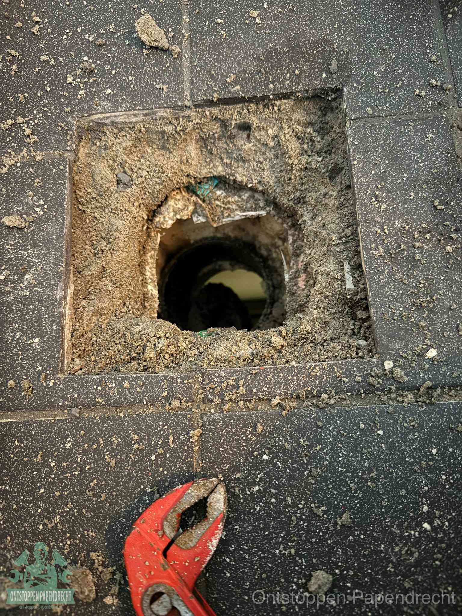 Een geopend rioolputje in een betontegelvloer met losgemaakt rooster en een zichtbare ronde afvoerbuis, het achterhalen waar de afvoerverstopping zit in Papendrecht