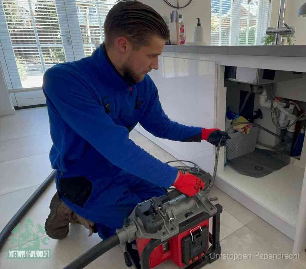 Een monteur met rode handschoenen knielt in een keuken en voert een elektrische ontstoppingsveer in tijdens het professioneel afvoer ontstoppen in Papendrecht